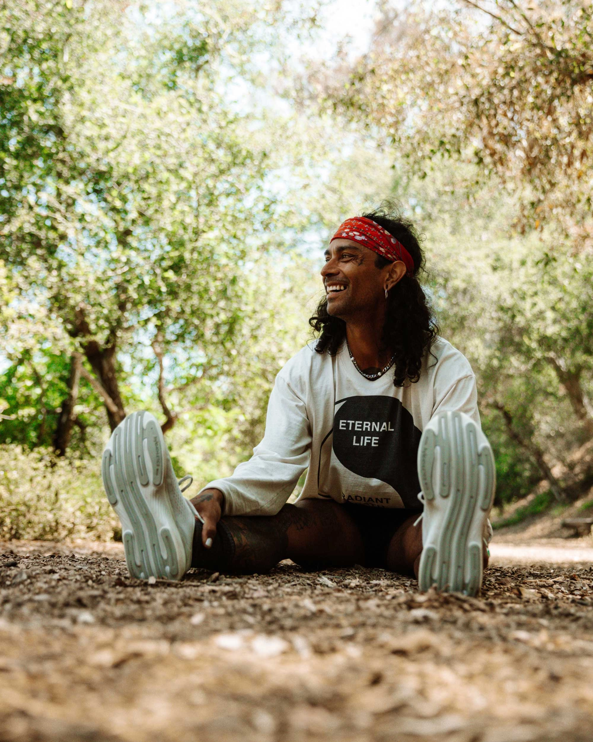 A runner in a red bandana sits laughing on a dirt path, soles of his hylo shoes to the camera