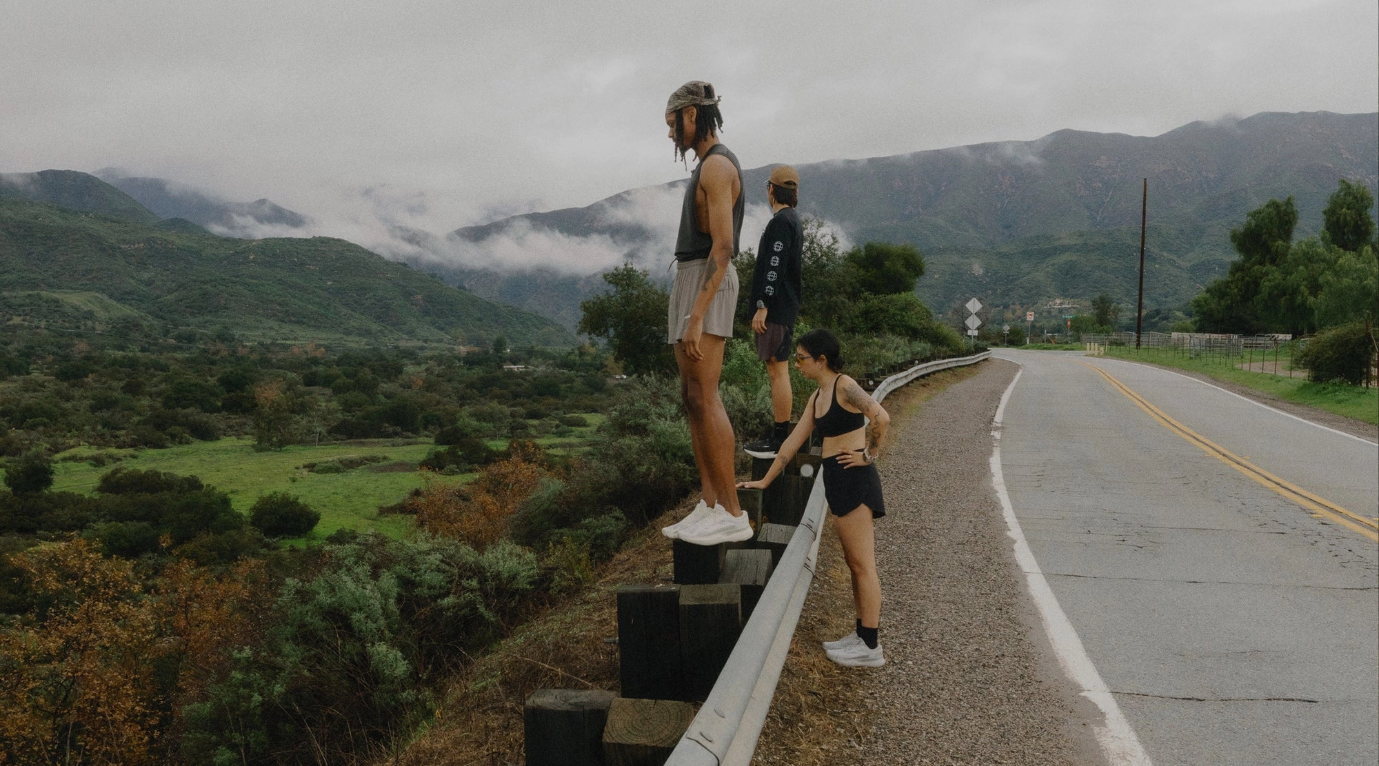 Two runners rest on a roadside barrier above a green valley